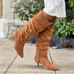 Brown extra wide suede knee-high boots worn outdoors on a stone pavement with greenery in the background.